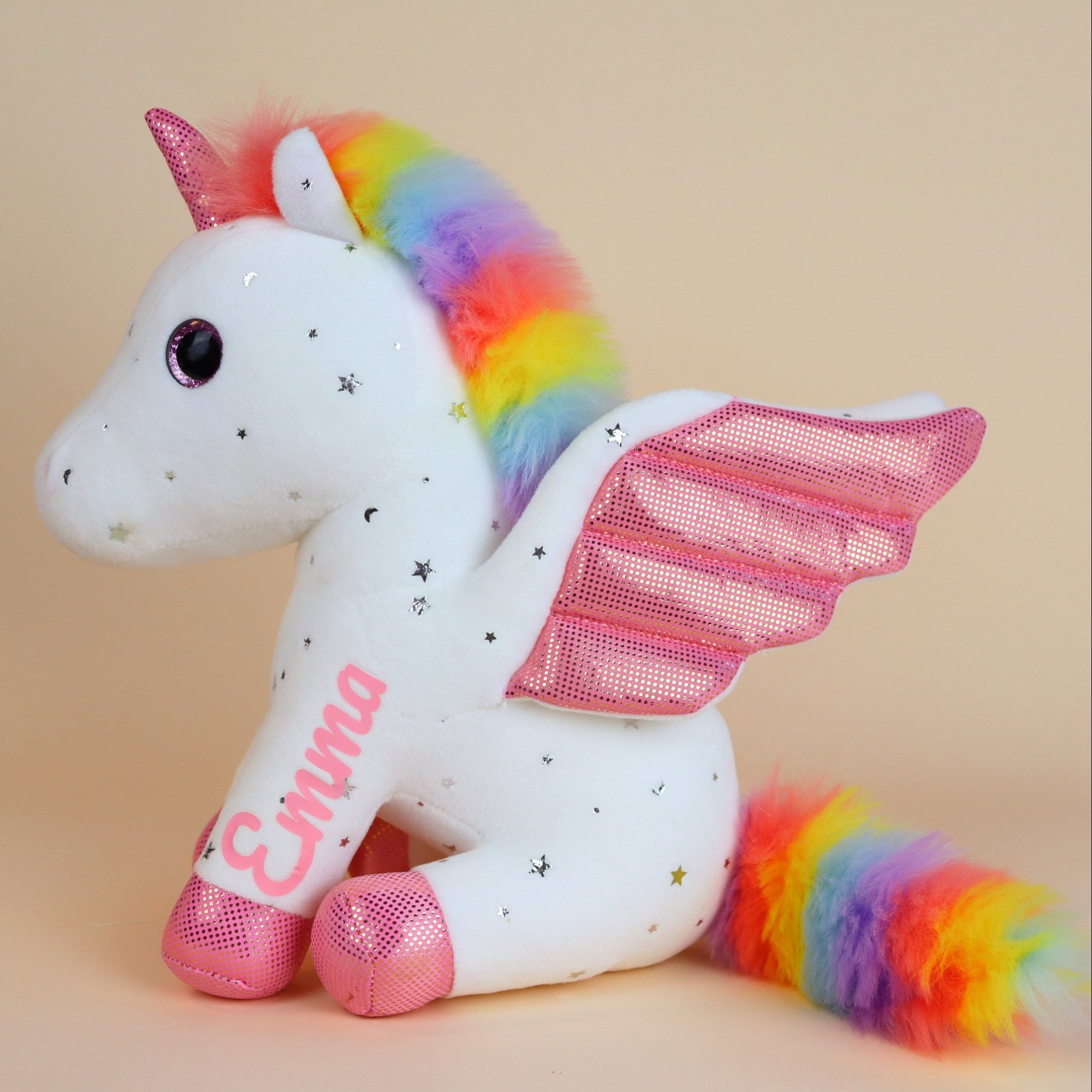 Stuffed unicorn toy with rainbow hair and tail, pink wings, and 'Emma' on a beige background